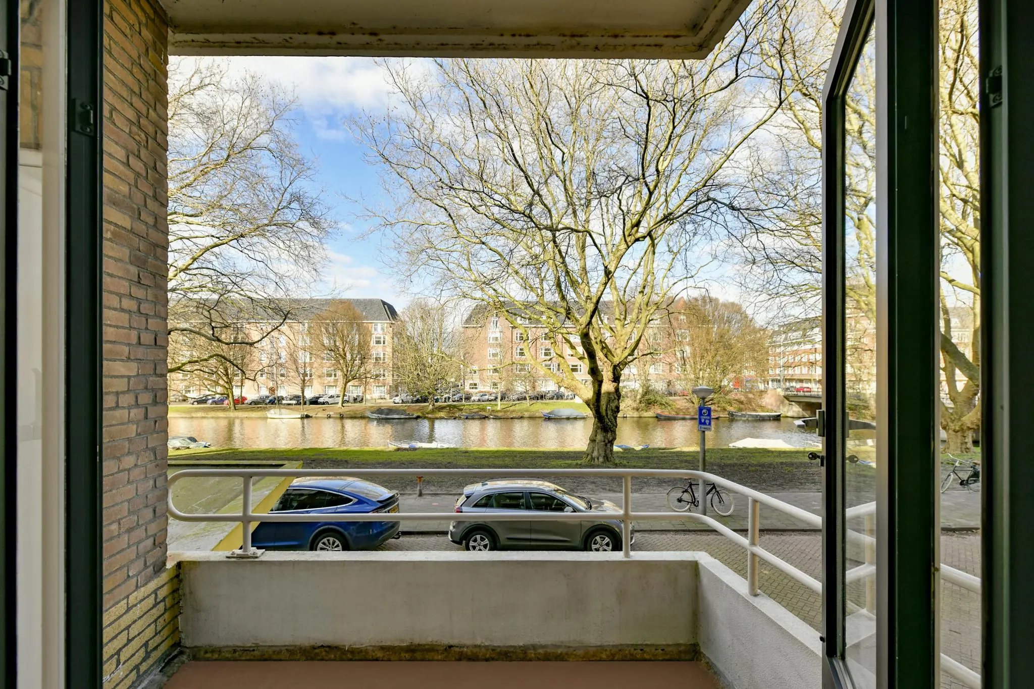 Uitzicht vanaf een balkon aan de Cornelis Dopperkade op een gracht met geparkeerde auto's, bomen en tegenoverliggende woongebouwen.